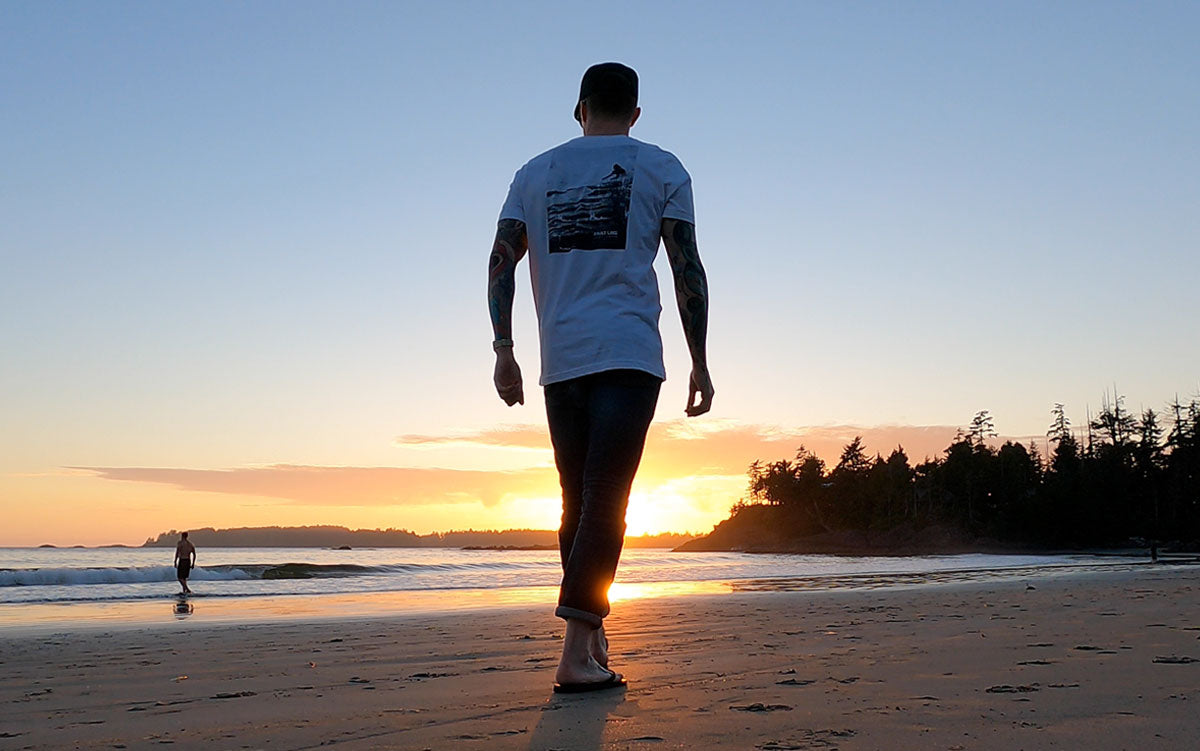 Man wearing Fault Line's Signature t-shirt walking along a beach at sunset